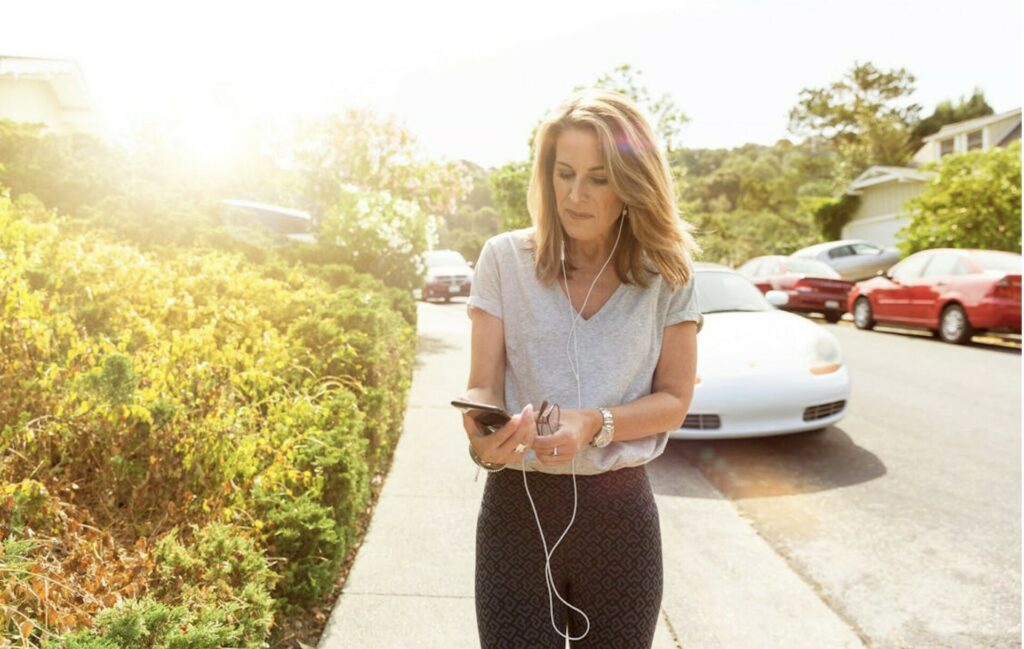 Femme marchant dans la rue avec des écouteurs, concentrée sur son téléphone, en pleine écoute ou enregistrement d’un message vocal.